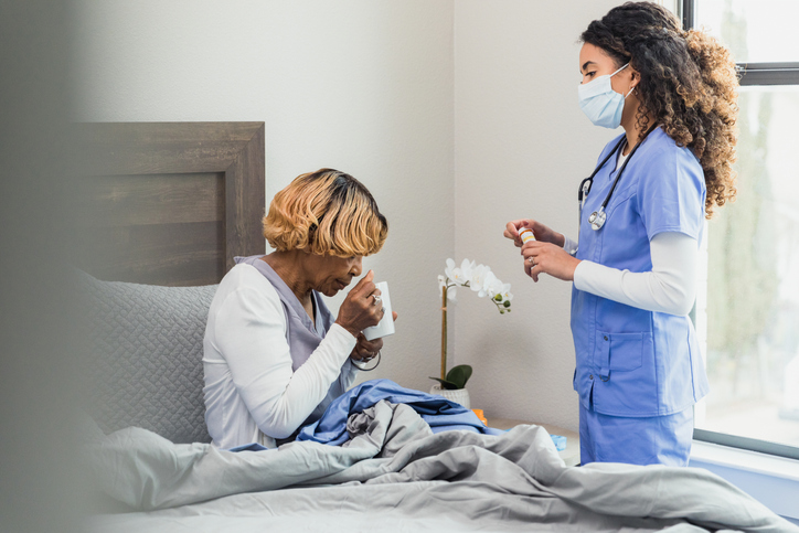 As nurse stands by bed, the senior woman takes pills