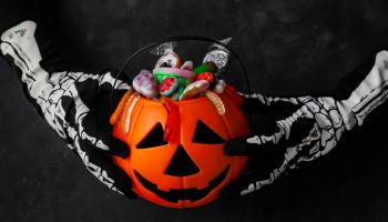 Jack O' Lantern, trick or treat bucket, filled with candies held by a skeleton hands on black background. Halloween celebration concept.