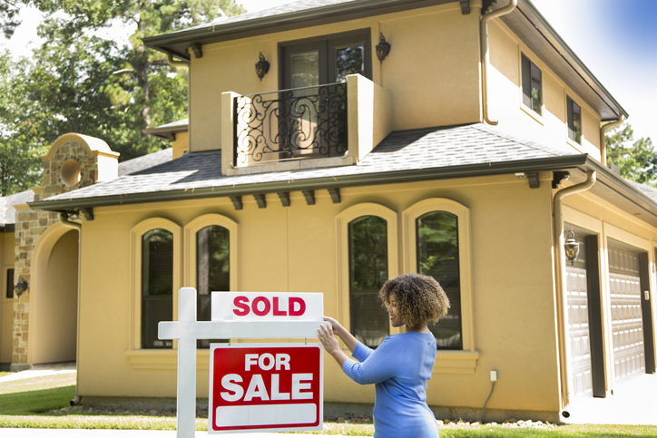 Lovely young adult realtor standing beside her for sale sign in front yard of home. She adds the SOLD sign and wears a blue top and jeans.