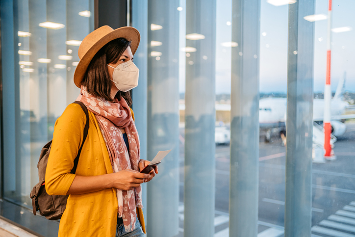 Young woman with mask looking the airport runway from the window