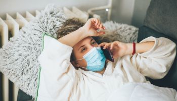 Sick woman with face protective mask using mobile phone and lying in bed