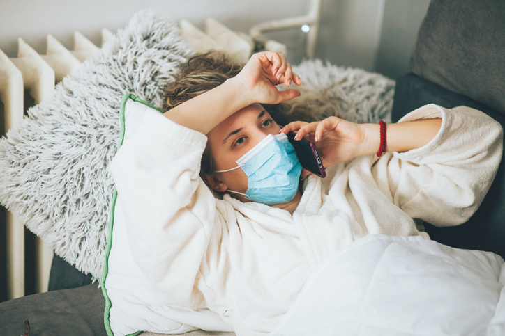 Sick woman with face protective mask using mobile phone and lying in bed