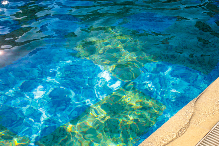 Close-up turquoise swimming pool water ripples with shimmering sunlight reflections beside a smooth tan tiled pool edge