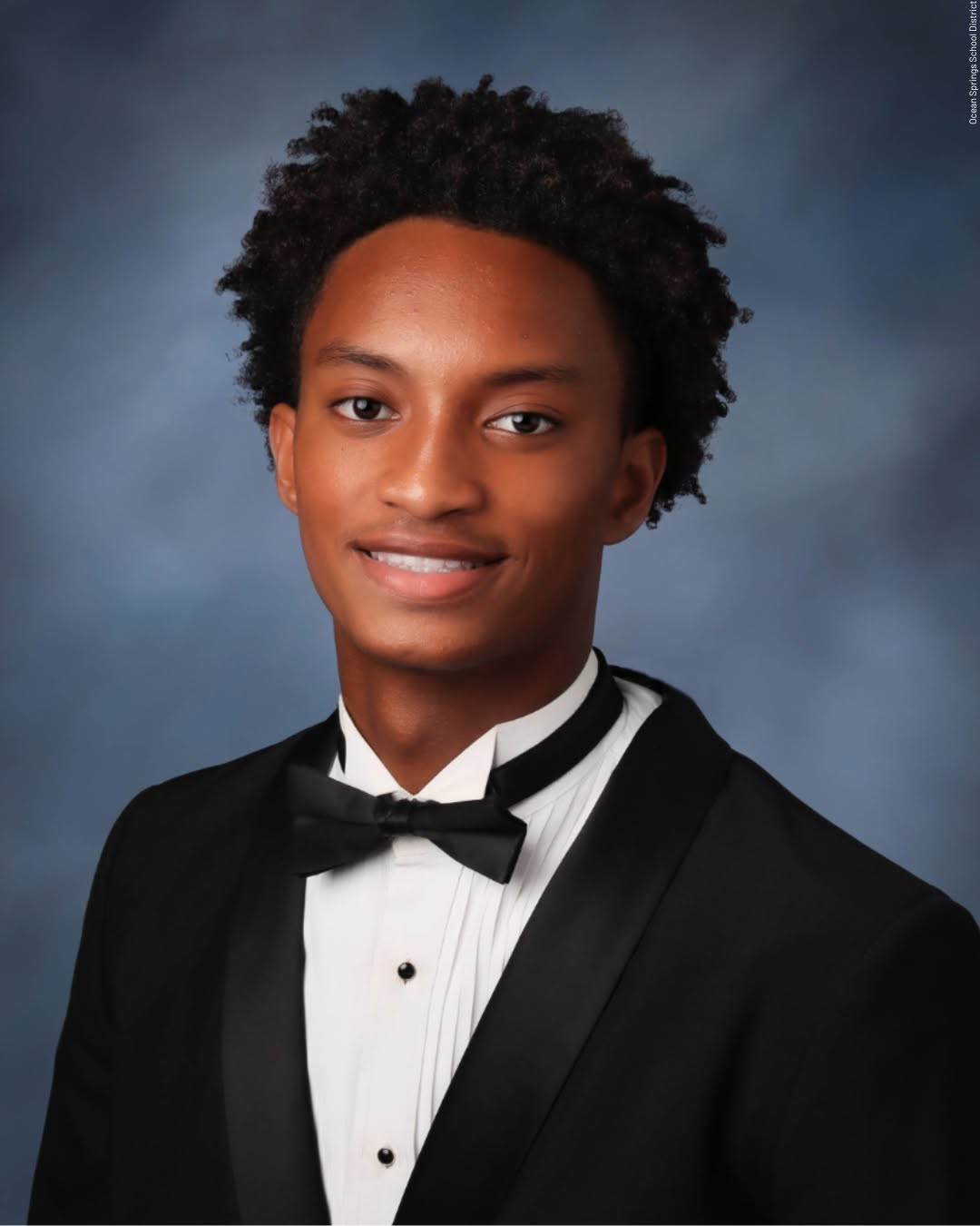 A young man in a tuxedo with a bow tie, smiling at the camera against a dark blue background.